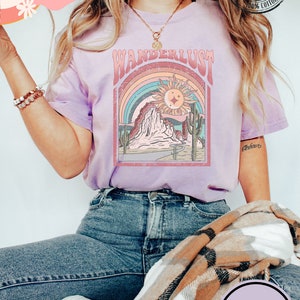 May include: A light purple t-shirt with a retro "Wanderlust" graphic featuring a desert landscape, rainbow, and sun. The shirt is made of 100% cotton and has a Comfort Colors tag. The word "ORCHID" is printed on a purple circle.