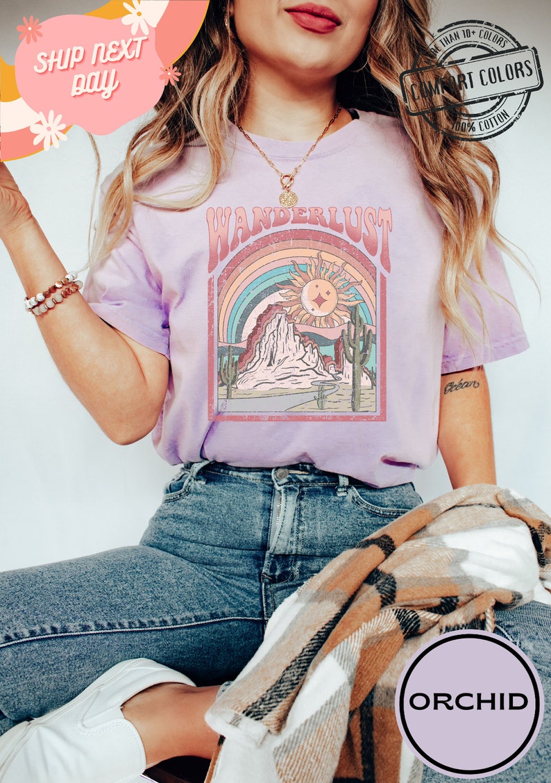 May include: A light purple t-shirt with a retro "Wanderlust" graphic featuring a desert landscape, rainbow, and sun. The shirt is made of 100% cotton and has a Comfort Colors tag. The word "ORCHID" is printed on a purple circle.