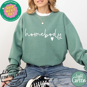 Może przedstawiać: Jasnozielona bluza z napisem "homebody" w białych literach i małym białym sercem. Bluza jest wykonana w 100% z bawełny i dostępna w r&oacute;żnych kolorach.