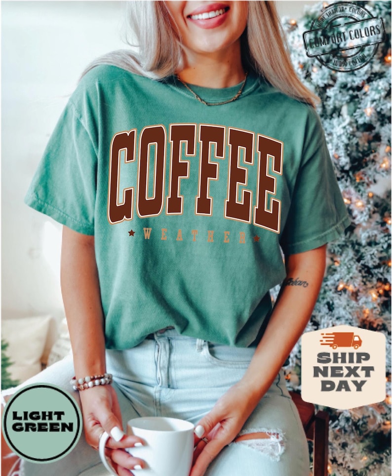 May include: A light green t-shirt with the text "COFFEE WEATHER" in brown and gold lettering. The shirt is being worn by a person.
