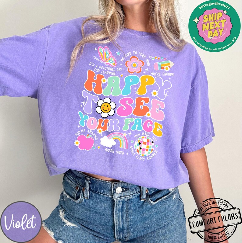 May include: Purple t-shirt with a colorful graphic that says "Happy to see your face". The graphic includes a rainbow, a disco ball, a flower, and an apple. The shirt is made of 100% cotton.