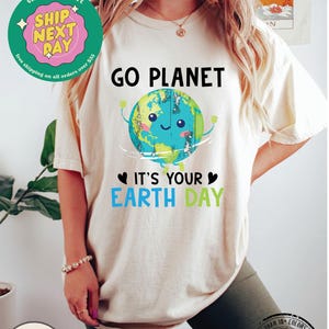 May include: A white t-shirt with a green, blue, and brown cartoon earth graphic. The text "GO PLANET" is above the earth and the text "IT'S YOUR EARTH DAY" is below the earth. The shirt is being worn by a person with long blonde hair.