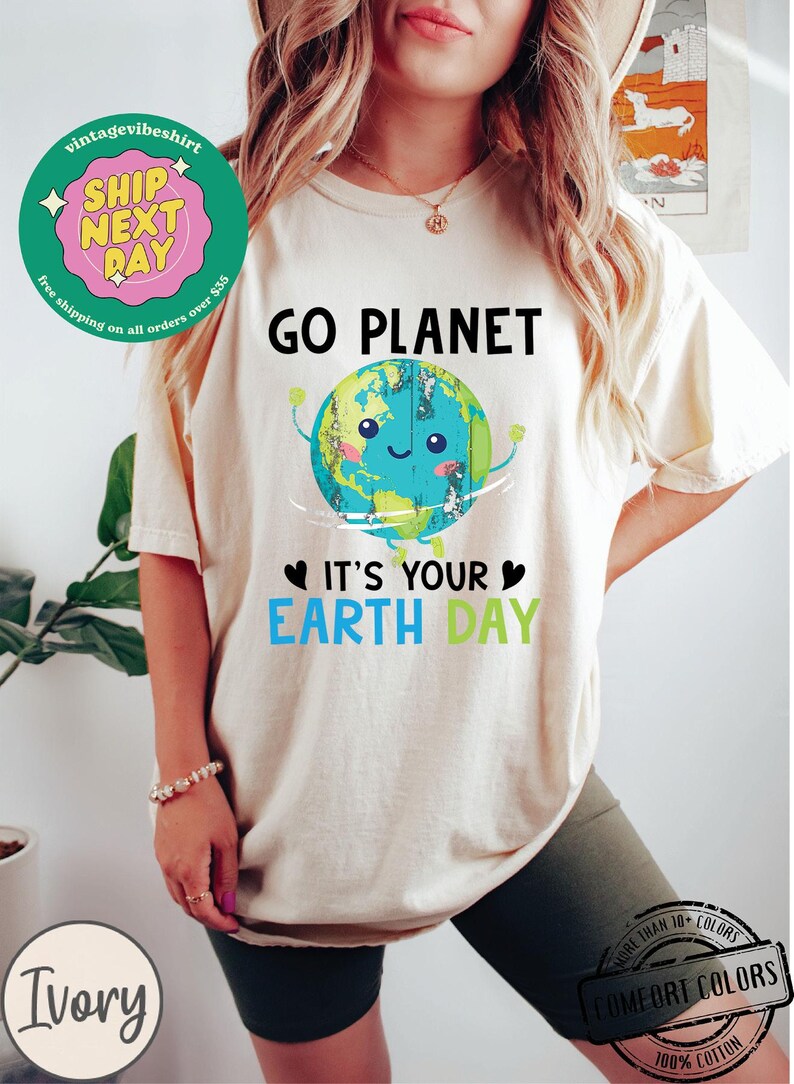 May include: A white t-shirt with a green, blue, and brown cartoon earth graphic. The text "GO PLANET" is above the earth and the text "IT'S YOUR EARTH DAY" is below the earth. The shirt is being worn by a person with long blonde hair.