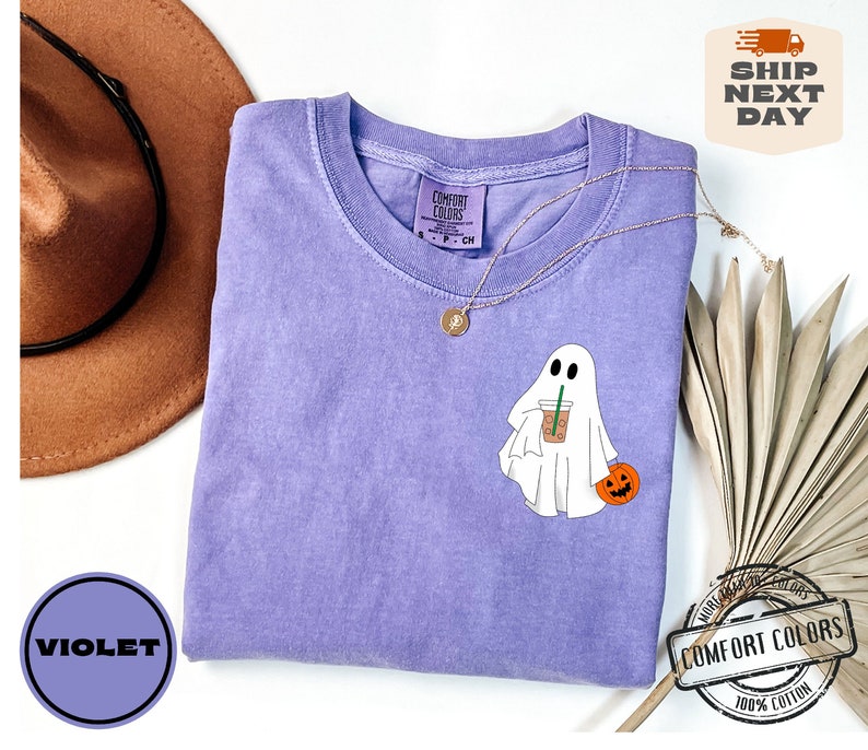 Peut inclure: Un t-shirt violet avec un fant&ocirc;me blanc tenant une citrouille et une tasse de caf&eacute;. Le t-shirt porte l'inscription "Comfort Colors" sur l'&eacute;tiquette.