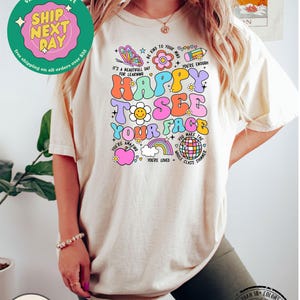 May include: A light beige t-shirt with a colorful graphic design featuring the text "Happy to see your face". The graphic includes a butterfly, a flower, a rainbow, a smiley face, and a disco ball. The shirt is made of 100% cotton.