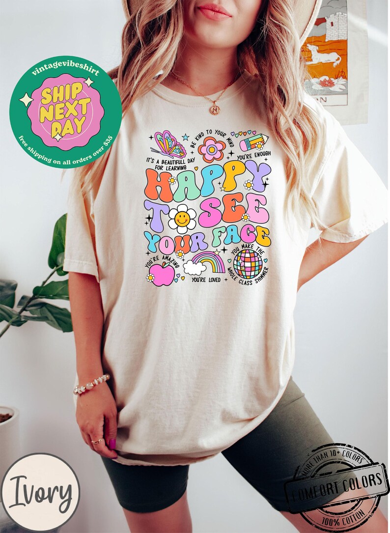 May include: A light beige t-shirt with a colorful graphic design featuring the text "Happy to see your face". The graphic includes a butterfly, a flower, a rainbow, a smiley face, and a disco ball. The shirt is made of 100% cotton.