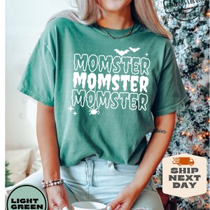 May include: A light green t-shirt with the text "Momster" repeated three times in white. The text is in a dripping font style. There are small white bats and stars above the text and a spider below the text.