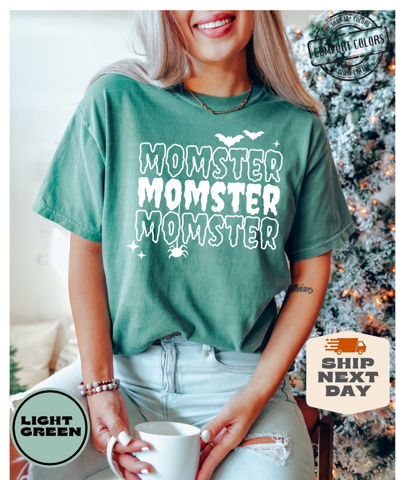 May include: A light green t-shirt with the text "Momster" repeated three times in white. The text is in a dripping font style. There are small white bats and stars above the text and a spider below the text.