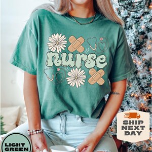 May include: A light green t-shirt with the word "nurse" in a retro font. The shirt is decorated with white daisies, red hearts, and plasters.
