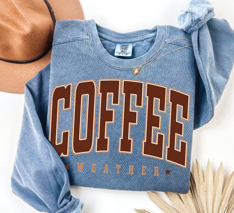May include: A blue sweatshirt with the text "COFFEE WEATHER" in brown letters with a white outline. The sweatshirt has a distressed look.
