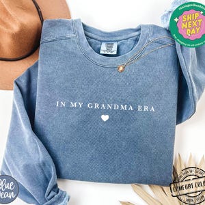 May include: A blue sweatshirt with the text "IN MY GRANDMA ERA" and a small white heart in the center.