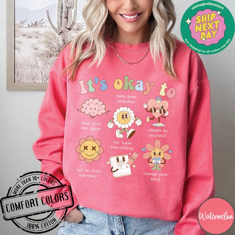May include: Pink sweatshirt with the text "It's okay to" and colorful illustrations of a brain, flowers, and a heart. The illustrations have text below them, such as "make some mistakes", "need your own space", and "always be yourself".