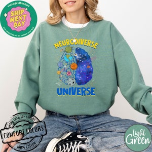 Op de afbeelding: Een lichtgroen sweatshirt met de tekst "Neurodiverse Universe" bedrukt op de voorkant. De tekst is in blauw en geel, en er is een afbeelding van een brein met planeten en sterren erin. Het sweatshirt is gemaakt van 100% katoen en is verkrijgbaar in verschillende kleuren.