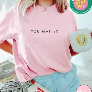 May include: A pink t-shirt with the text "YOU MATTER" printed in black on the front. The shirt is being worn by a person with blue shorts and a gold necklace.