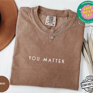 May include: A brown t-shirt with the text "YOU MATTER" printed in white on the front. The shirt is made of 100% cotton and is available in a variety of sizes.