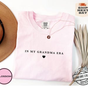 May include: Pink t-shirt with the text "IN MY GRANDMA ERA" and a small black heart. The shirt is made of 100% cotton.