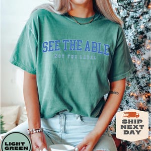 May include: A light green t-shirt with the text "See the Able, Not the Label" in blue lettering. The shirt is worn by a person with a gold necklace and a light blue denim skirt.