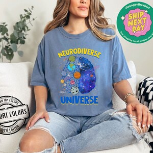 Op de afbeelding: Een blauw t-shirt met een afbeelding van een brein met planeten en sterren erin. De tekst "Neurodiverse Universe" staat op het t-shirt gedrukt.