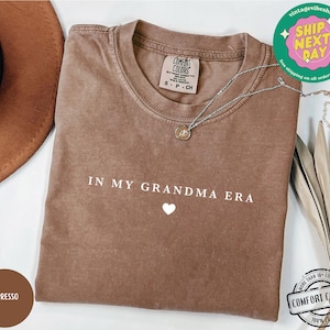 May include: A brown t-shirt with the text "IN MY GRANDMA ERA" in white lettering and a small white heart below the text. The shirt is made of 100% cotton.
