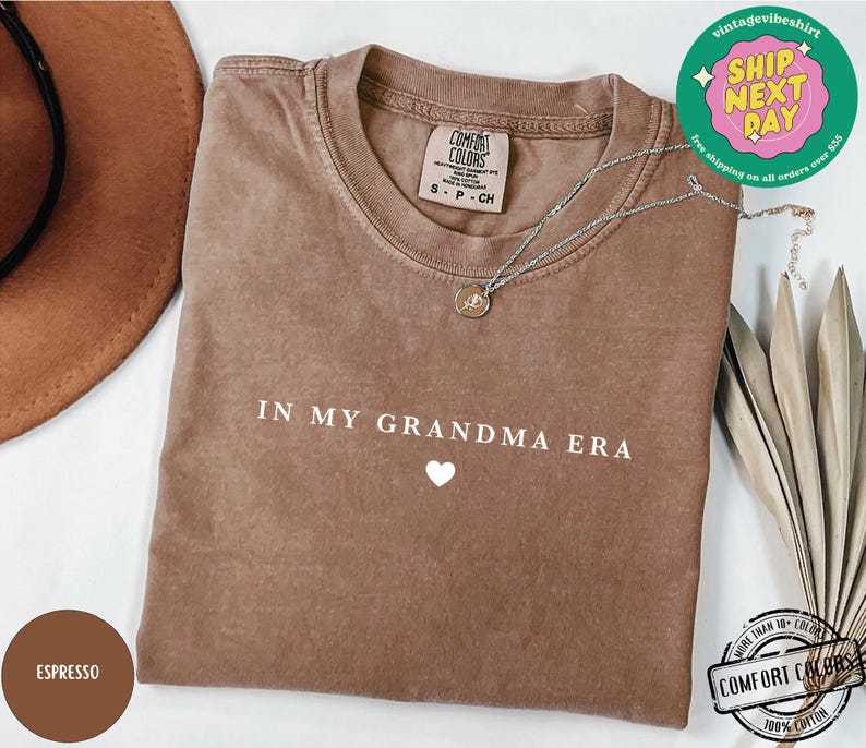 May include: A brown t-shirt with the text "IN MY GRANDMA ERA" in white lettering and a small white heart below the text. The shirt is made of 100% cotton.