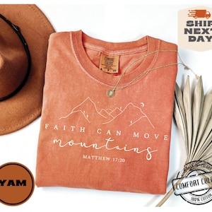 May include: A coral-colored t-shirt with a white graphic of mountains and the text "FAITH CAN MOVE mountains MATTHEW 17:20".