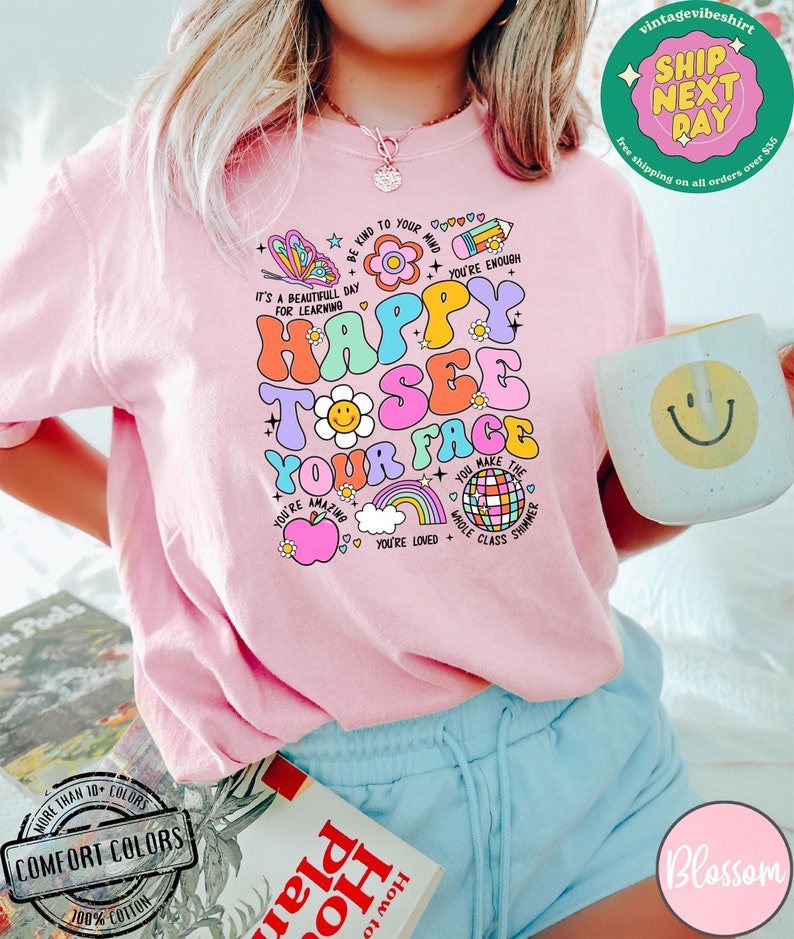 May include: Pink t-shirt with a colorful graphic design that says "Happy to see your face". The graphic includes a butterfly, flower, rainbow, apple, cloud, and disco ball. The shirt is made of 100% cotton.