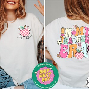 K&ouml;nnte beinhalten: Wei&szlig;es T-Shirt mit einer pinkfarbenen und gr&uuml;nen Grafik, auf der "In My Teacher Era" mit einem roten Apfel, einem Bleistift und Sternen steht. Die Vorderseite des Shirts hat einen pinkfarbenen und wei&szlig;en karierten Apfel mit einem gr&uuml;nen Stiel und Bl&auml;ttern. Der Text "Miss Olivia" steht unter dem Apfel.