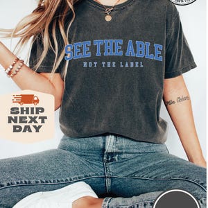 May include: A dark grey t-shirt with the text "See the Able, Not the Label" in blue lettering. The shirt is made of 100% cotton and is available in over 10 colours.