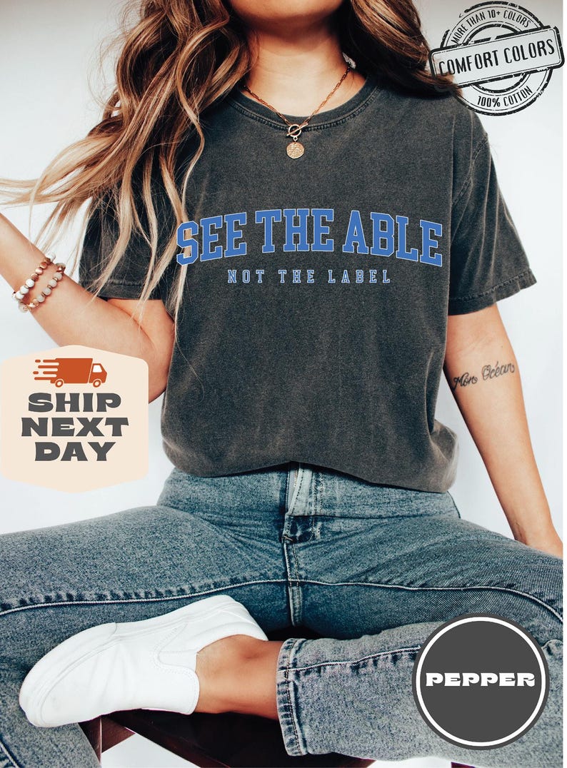 May include: A dark grey t-shirt with the text "See the Able, Not the Label" in blue lettering. The shirt is made of 100% cotton and is available in over 10 colours.