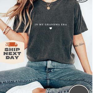 May include: A dark gray t-shirt with the text "IN MY GRANDMA ERA" and a small white heart. The shirt is made of 100% cotton and is available in a variety of colors.