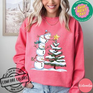 以下が含まれることがあります： クリスマスのデザインが施されたピンクのトレーナー。3人の雪だるまが積み重なっており、一番上の雪だるまが緑色のクリスマスツリーの頂上にある星に手を伸ばしています。