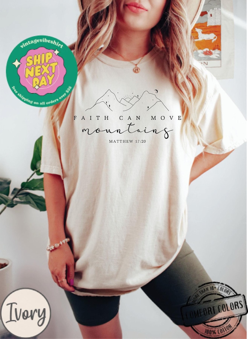 May include: Ivory t-shirt with a black graphic of mountains, a sun, and a moon. The text "FAITH CAN MOVE mountains" and "MATTHEW 17:20" is printed below the graphic.