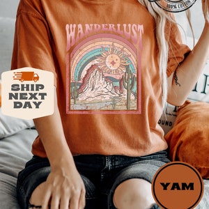 May include: Burnt orange t-shirt with a graphic featuring the word "WANDERLUST" above a desert landscape with a rainbow, sun, and cacti. The shirt is made of 100% cotton and has a "COMFORT COLORS" label.