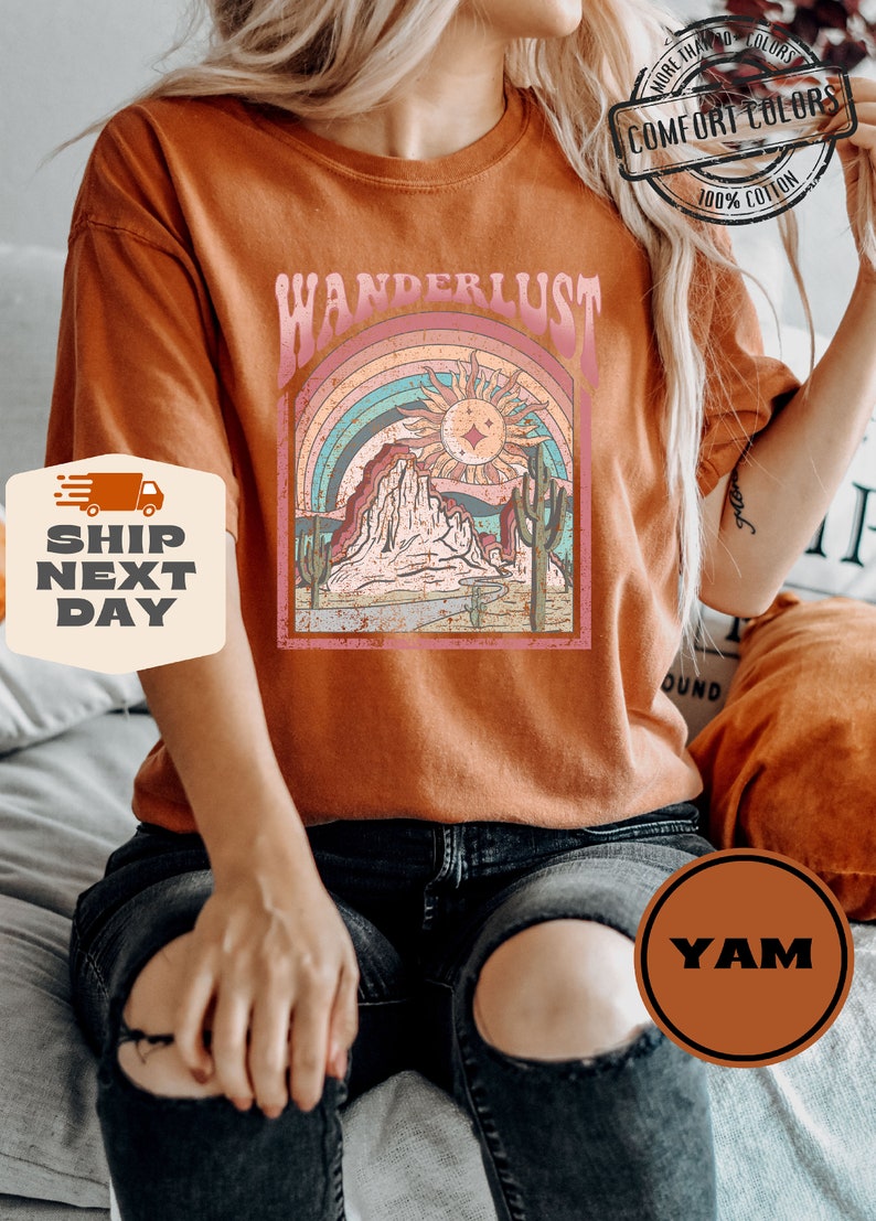 May include: Burnt orange t-shirt with a graphic featuring the word "WANDERLUST" above a desert landscape with a rainbow, sun, and cacti. The shirt is made of 100% cotton and has a "COMFORT COLORS" label.