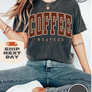 May include: A dark gray t-shirt with the text "COFFEE WEATHER" in brown letters. The shirt is worn by a person with long brown hair and blue jeans.