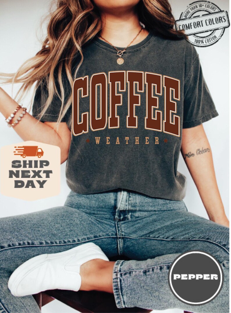 May include: A dark gray t-shirt with the text "COFFEE WEATHER" in brown letters. The shirt is worn by a person with long brown hair and blue jeans.