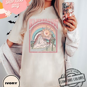 May include: Ivory-colored t-shirt with a retro graphic design featuring the word "WANDERLUST" above a desert landscape with a rainbow, sun, and cacti. The shirt is made of 100% cotton and is part of the Comfort Colors collection.