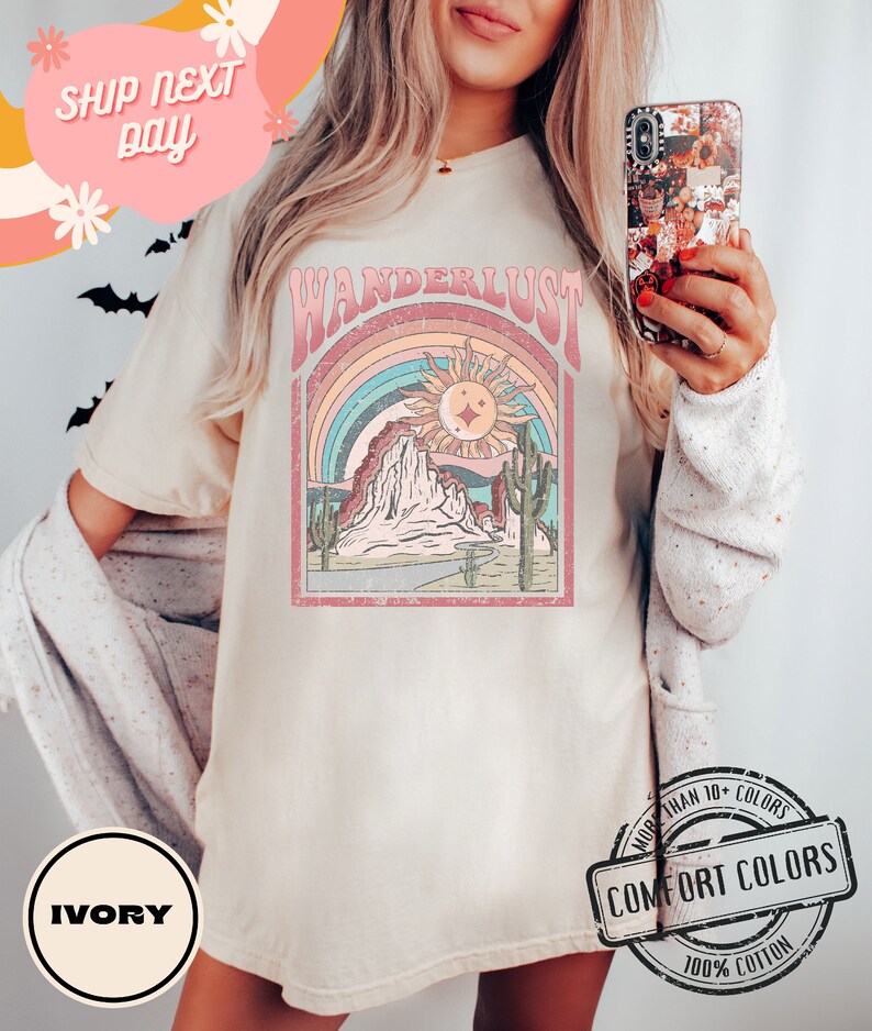 May include: Ivory-colored t-shirt with a retro graphic design featuring the word "WANDERLUST" above a desert landscape with a rainbow, sun, and cacti. The shirt is made of 100% cotton and is part of the Comfort Colors collection.