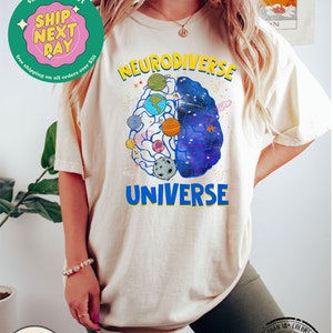 Op de afbeelding: Een cr&egrave;mekleurig T-shirt met een afbeelding van een brein met planeten en sterren erin. De tekst "Neurodiverse Universe" is op het shirt gedrukt.