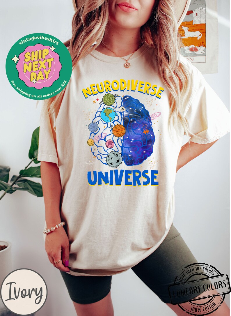 Op de afbeelding: Een cr&egrave;mekleurig T-shirt met een afbeelding van een brein met planeten en sterren erin. De tekst "Neurodiverse Universe" is op het shirt gedrukt.
