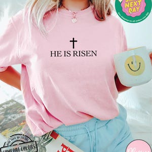 May include: A pink t-shirt with the text "HE IS RISEN" and a cross above it. The shirt is being worn by a person with long blonde hair.