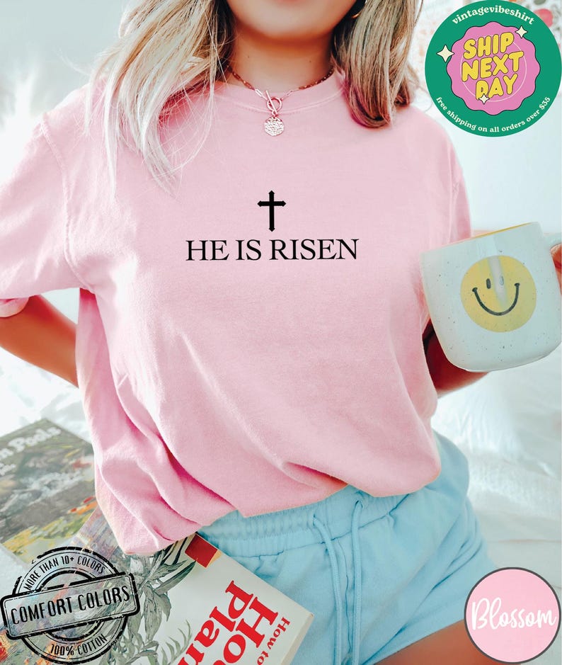 May include: A pink t-shirt with the text "HE IS RISEN" and a cross above it. The shirt is being worn by a person with long blonde hair.