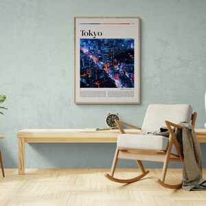 Może przedstawiać: Ramka z plakatem przedstawiającym zdjęcie nocnego panoramy miasta. Plakat zatytułowany jest "Tokyo" i ma białe tło z czarnym tekstem. Plakat wisi na jasnoniebieskiej ścianie nad drewnianym fotelem bujanym.