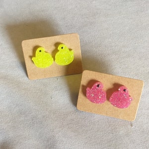 May include: Two pairs of glittery chick-shaped stud earrings. One pair is bright yellow, the other is pink. Each pair is mounted on a small, rectangular cardboard backing. The earrings are set against a neutral, textured background.