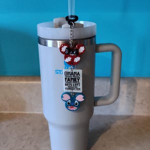 Puede incluir: Un vaso de acero inoxidable gris con tapa y asa negras. Una pajita se inserta a trav&eacute;s de la tapa. Un amuleto con una cabeza de Minnie Mouse roja y blanca, una cabeza de Stitch azul y una etiqueta blanca con el texto "Ohana means family. No one gets left behind or forgotten" est&aacute; unido a la tapa.