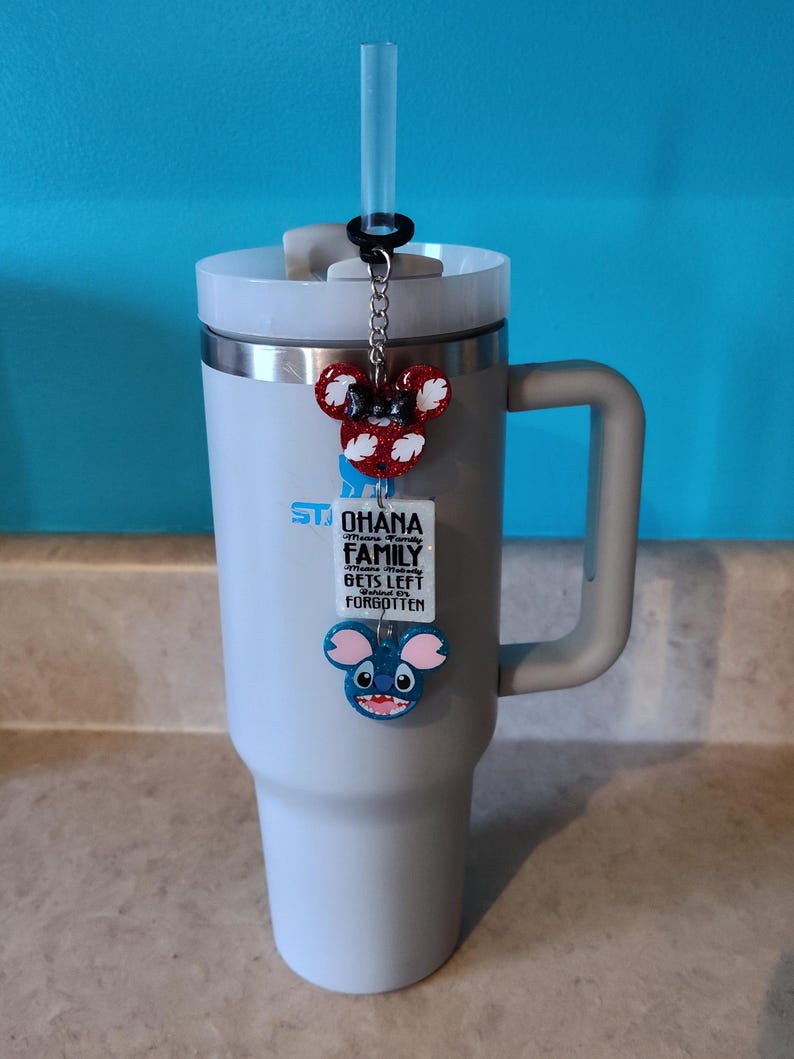 Puede incluir: Un vaso de acero inoxidable gris con tapa y asa negras. Una pajita se inserta a trav&eacute;s de la tapa. Un amuleto con una cabeza de Minnie Mouse roja y blanca, una cabeza de Stitch azul y una etiqueta blanca con el texto "Ohana means family. No one gets left behind or forgotten" est&aacute; unido a la tapa.