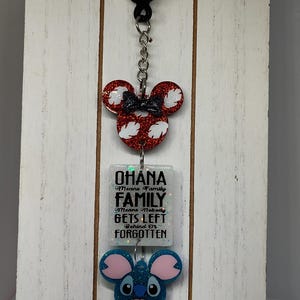 Puede incluir: Un llavero blanco con un dije de cabeza de Mickey Mouse rojo y blanco, un dije cuadrado blanco con el texto "Ohana Means Family Family Means Nobody Gets Left Behind Or Forgotten", y un dije de cabeza de Stitch azul.