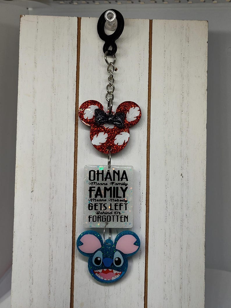 Puede incluir: Un llavero blanco con un dije de cabeza de Mickey Mouse rojo y blanco, un dije cuadrado blanco con el texto "Ohana Means Family Family Means Nobody Gets Left Behind Or Forgotten", y un dije de cabeza de Stitch azul.