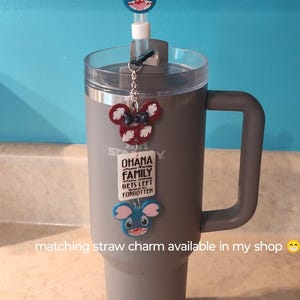 May include: A gray insulated tumbler with a handle, featuring a clear lid and a straw. Adorned with three cartoon character charms: a blue glittery character on the straw, a red and white charm, and a blue glittery charm at the bottom.