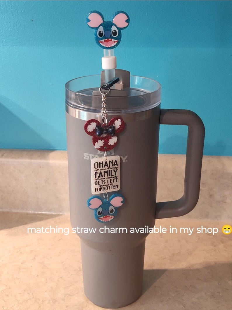 May include: A gray insulated tumbler with a handle, featuring a clear lid and a straw. Adorned with three cartoon character charms: a blue glittery character on the straw, a red and white charm, and a blue glittery charm at the bottom.